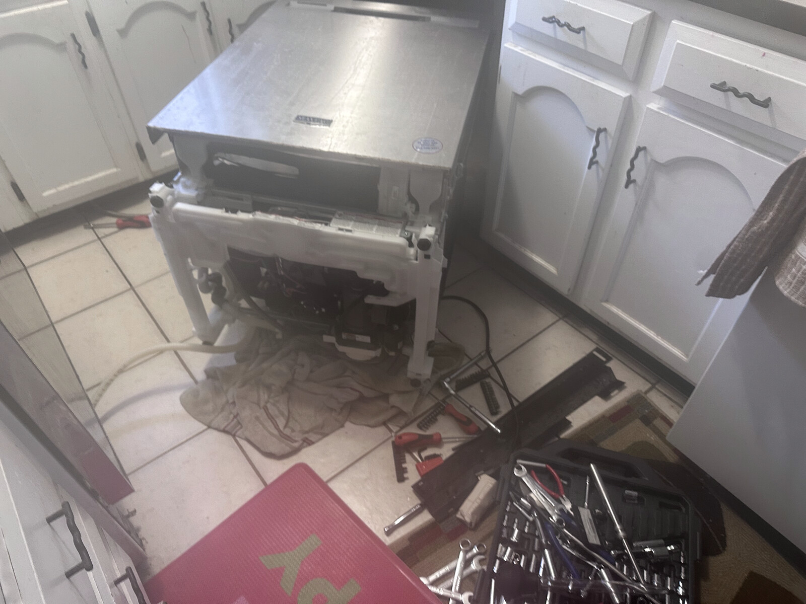 White-cabinet kitchen with a dishwasher pulled out from the cabinet and a technician's toolbox on the tile floor during a real repair