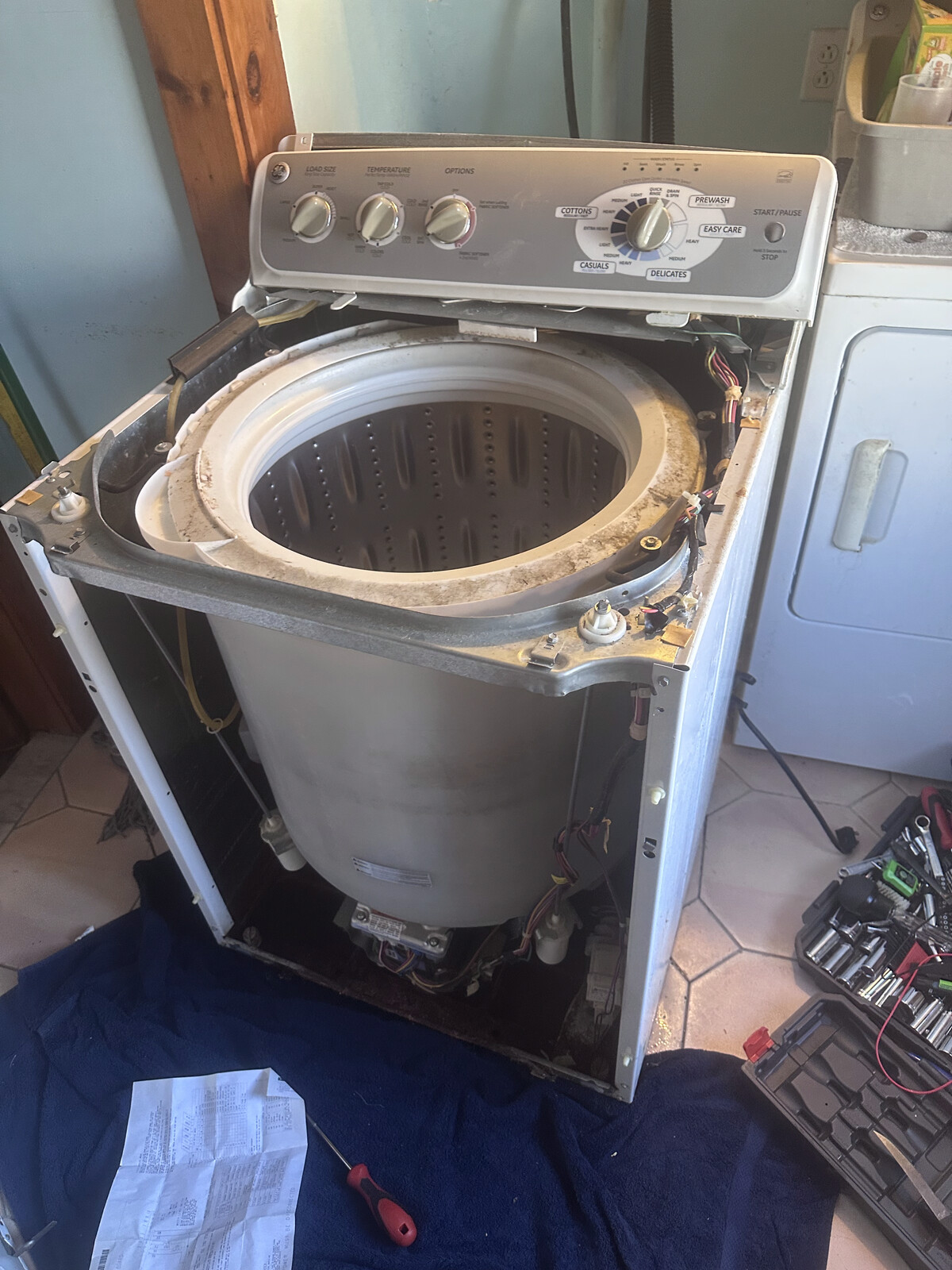 GE top-load washer with the outer cabinet opened and a technician's toolbox on the tile floor during a real repair