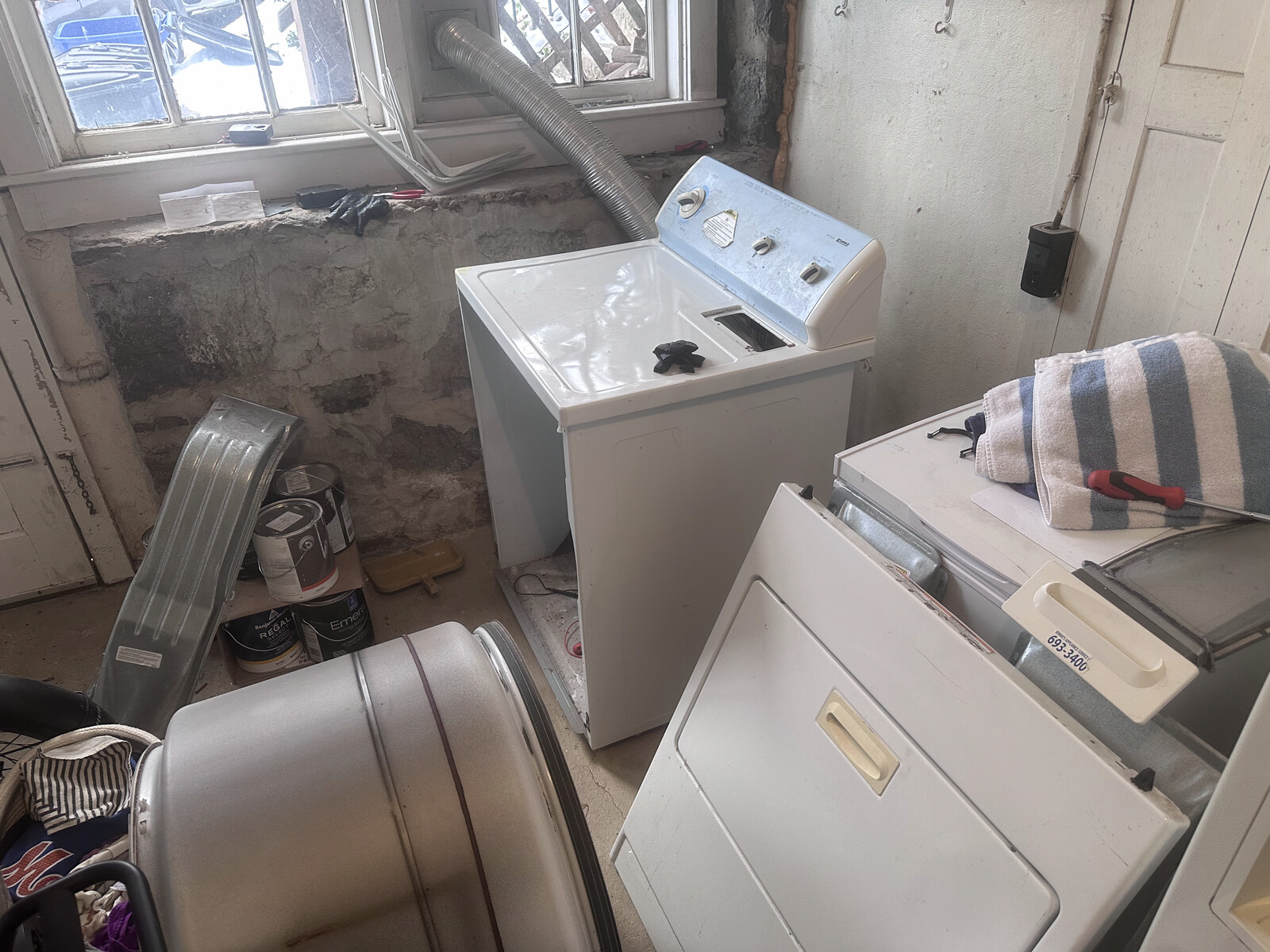 Older top-load washer and dryer pair in a basement laundry with a drum on the floor mid-repair
